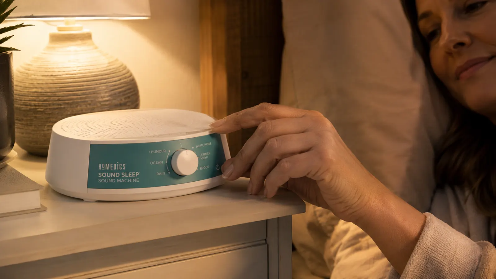 Hand adjusting the volume dial on the Homedics SoundSleep white noise machine on a bedside table