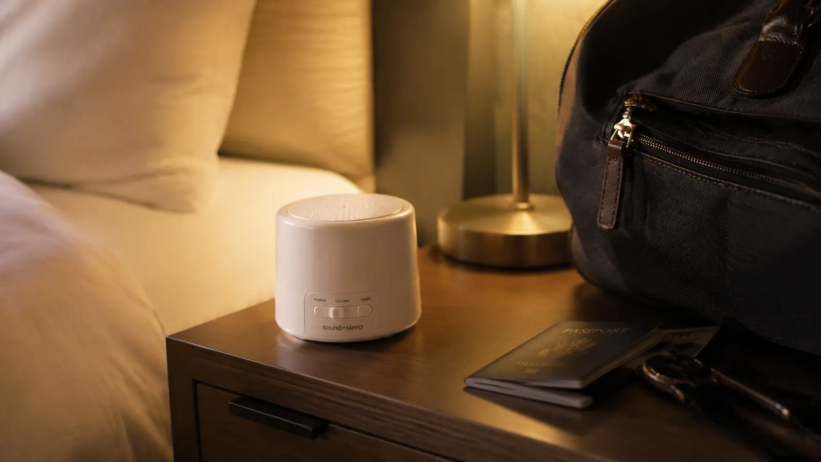 Compact white noise machine on a hotel nightstand beside a travel bag, suggesting portability