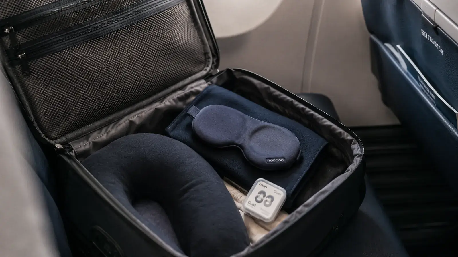 Travel scene showing a carry-on bag open with a sleep mask, earplugs, and a travel pillow inside